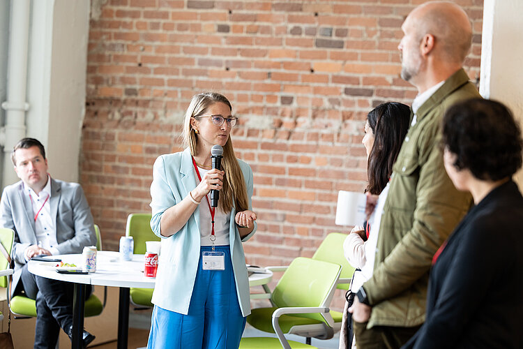 International Entrepreneurship Start-up Coach Vanessa hält ein Mikrofon in der Hand und spricht zu einer Gruppe von Entrepreneuren.
